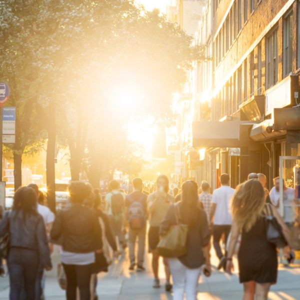 Man stands in the middle of a busy sidewalk looking at his cell phone while crowds of people walk around on 14th Street in Manhattan, New York City with the glow of sunlight in the background.